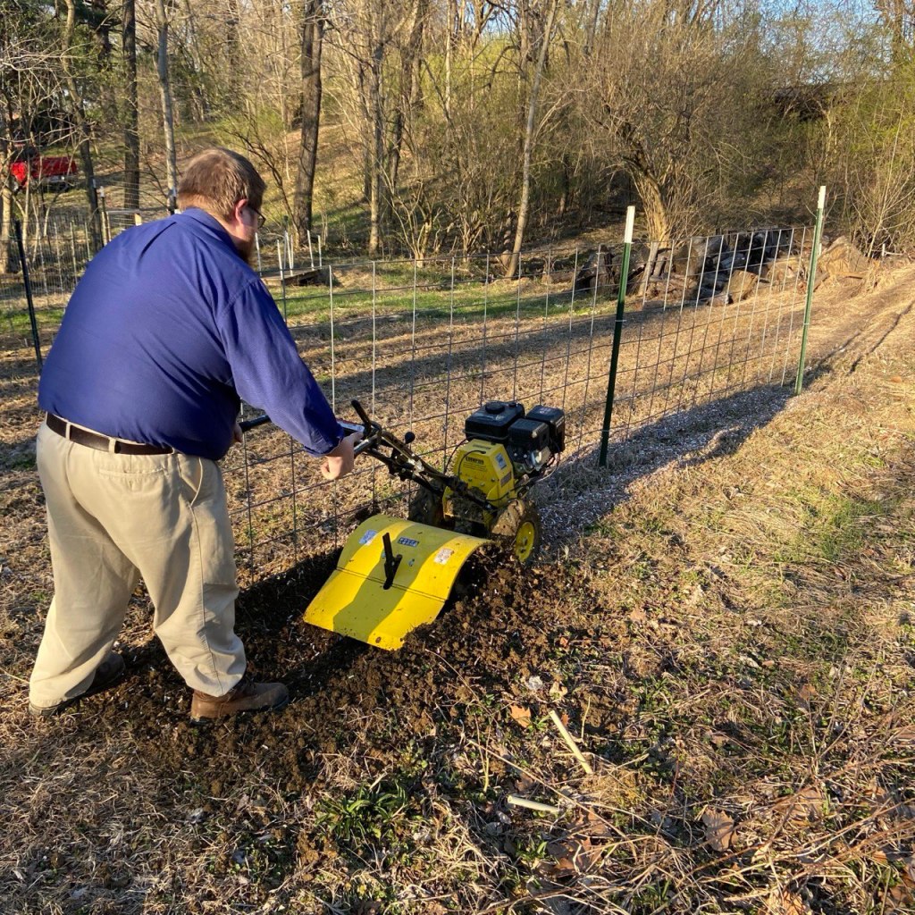 A man using a yellow rototiller to prepare a garden bed along a fence line.