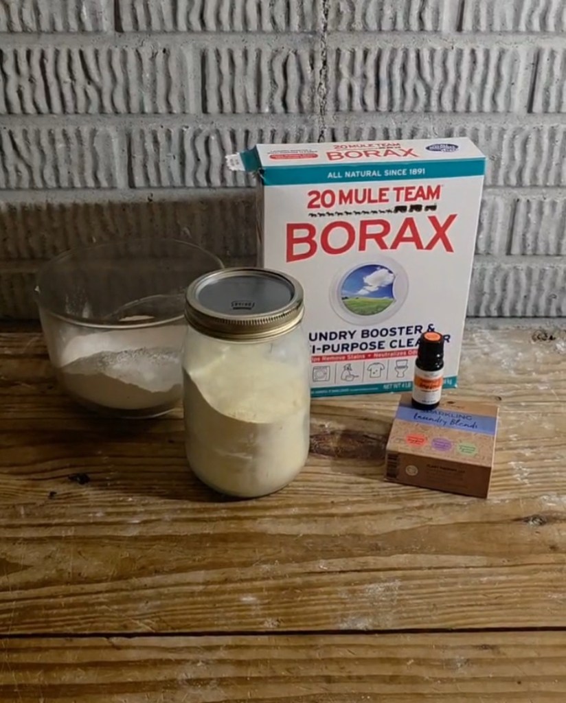 A box of Borax, a bar of Fels-Naptha, a box of essential oils, a bowl of washing soda, and a jar of laundry detergent powder. 